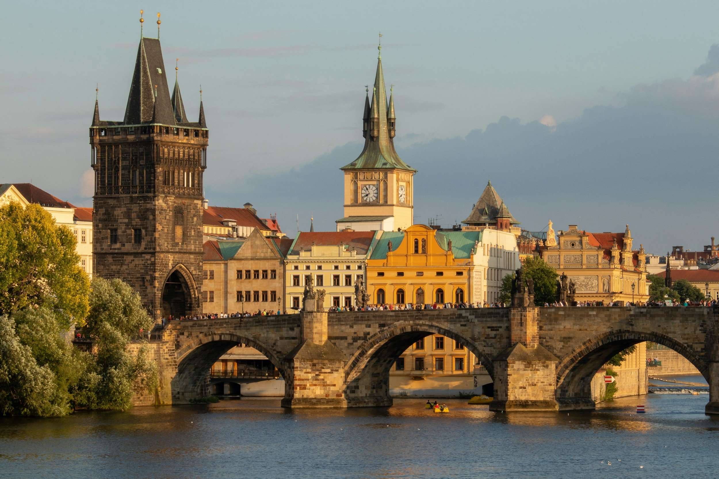 Historic stone bridge with arches over a river in Prague, featuring Gothic towers and Baroque buildings in the background at sunset.